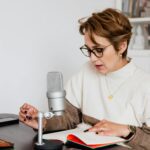 Female narrator in glasses reading out loud from book while sitting at desk with microphone and recording audiobook in cozy study